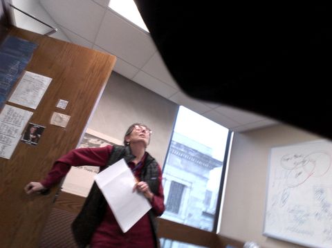 Person holding papers in an office or classroom with wooden paneling and windows.