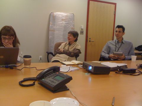 three people sit around a conference table with a laptop, projector, and phone in a meeting room