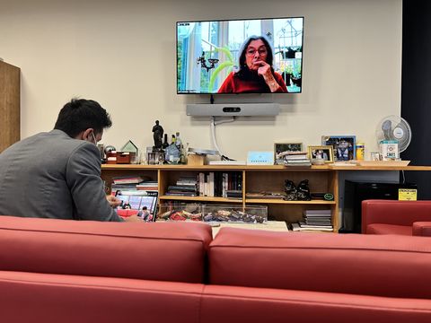a man sits on a red couch in an office lounge while a woman appears on a wall-mounted TV