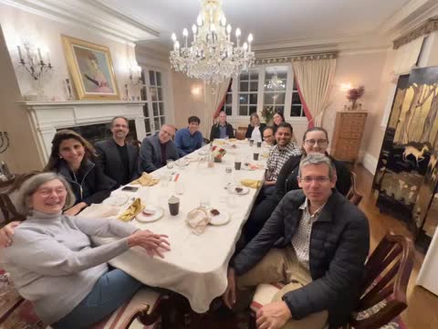 A group of people gathered around a formal dining table in an elegant room with a crystal chandelier and fireplace.