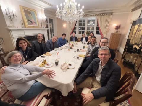 Group of people gathered around a dining table in an elegant room with a crystal chandelier and fireplace.