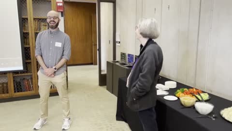 Two people in conversation at an indoor event with a refreshment table and bookshelf visible.