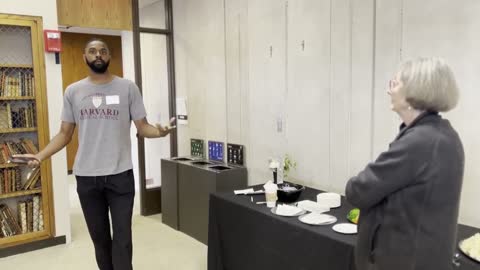 A man in a Harvard Medical School shirt presents to a woman at an indoor event with refreshments.