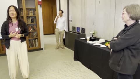 A woman presents to a small group in a library or office space with bookshelves and a refreshment table.