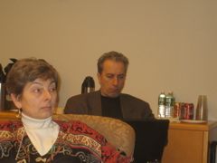 Two people seated indoors with beverages and items on a shelf behind them.